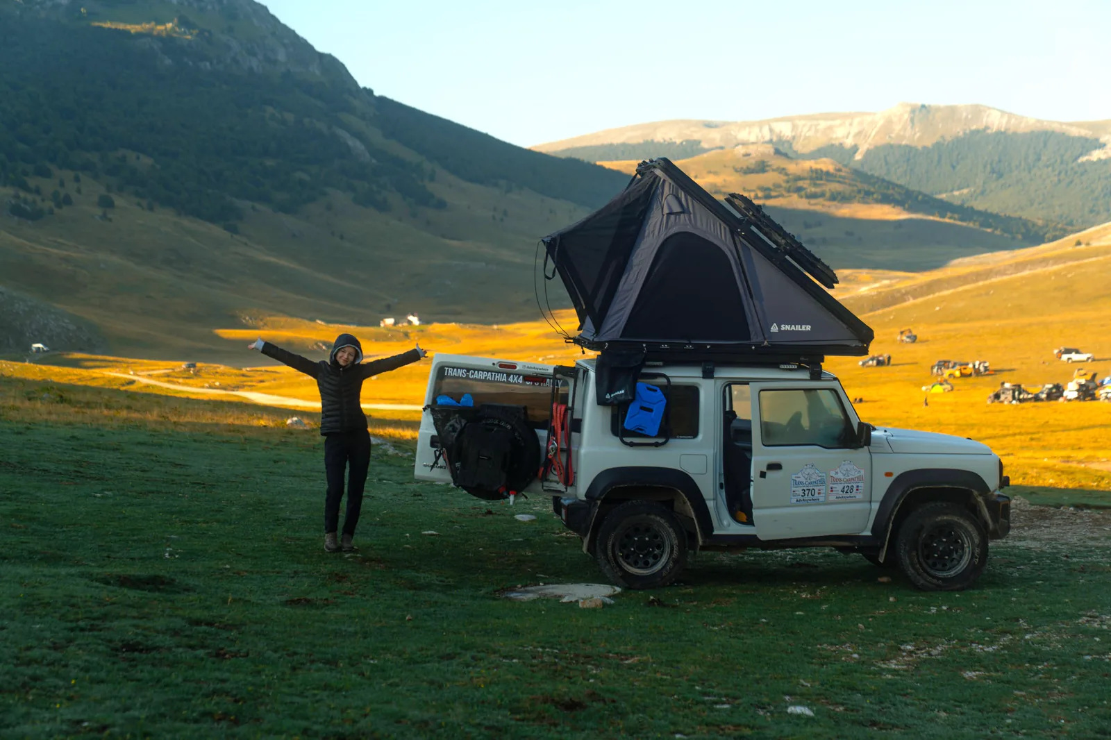 Traveller beside an off-road vehicle with an open SNAILER roof tent in a mountain valley